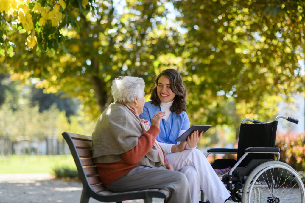 nurse and elderly woman sitting on bench in park during warm autumn day. Young caregiver looking through photos of senior patient's family on tablet